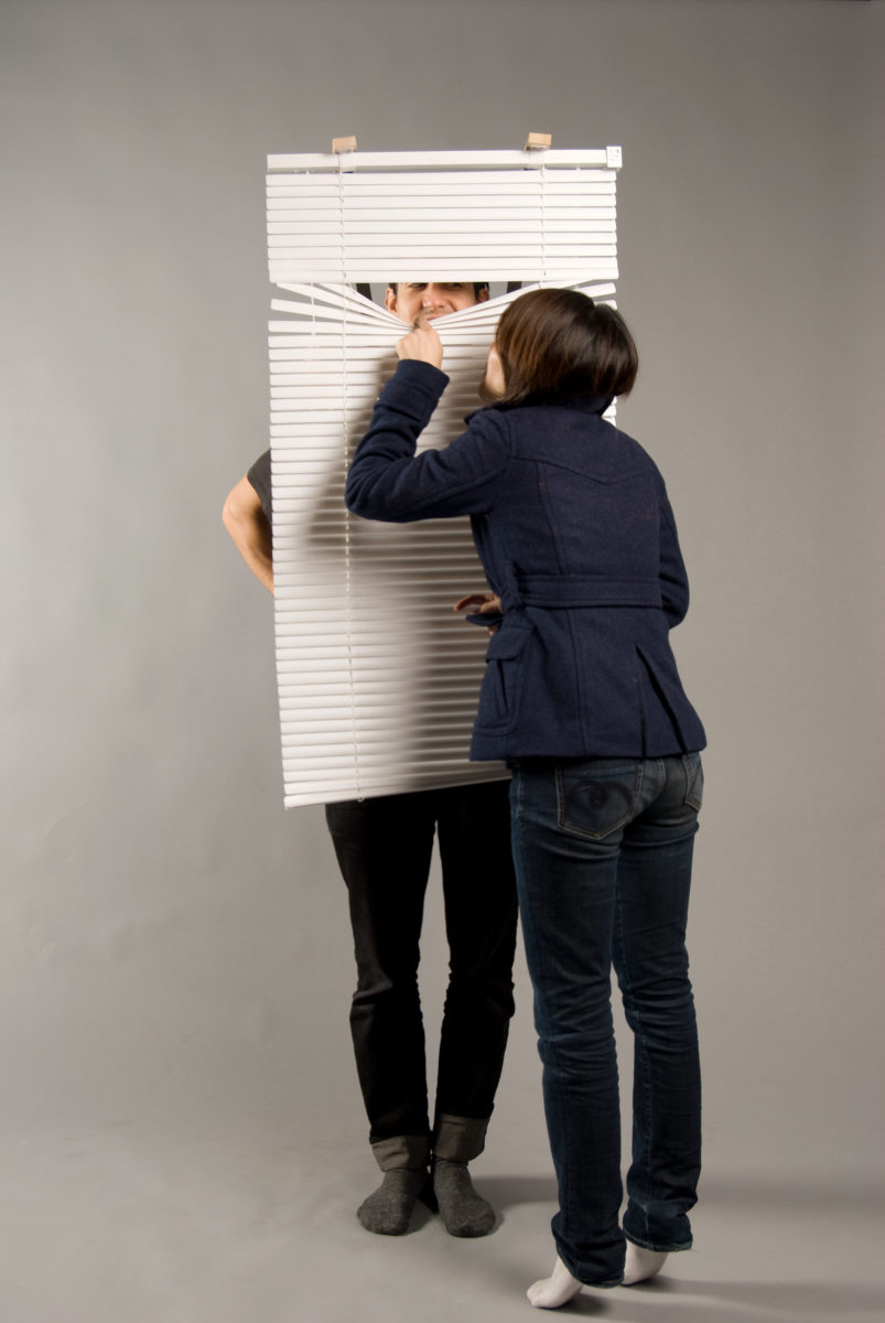 Man with venetian blinds hanging in front of his body; a woman in front of him is peaking through the blinds to see his part of his face